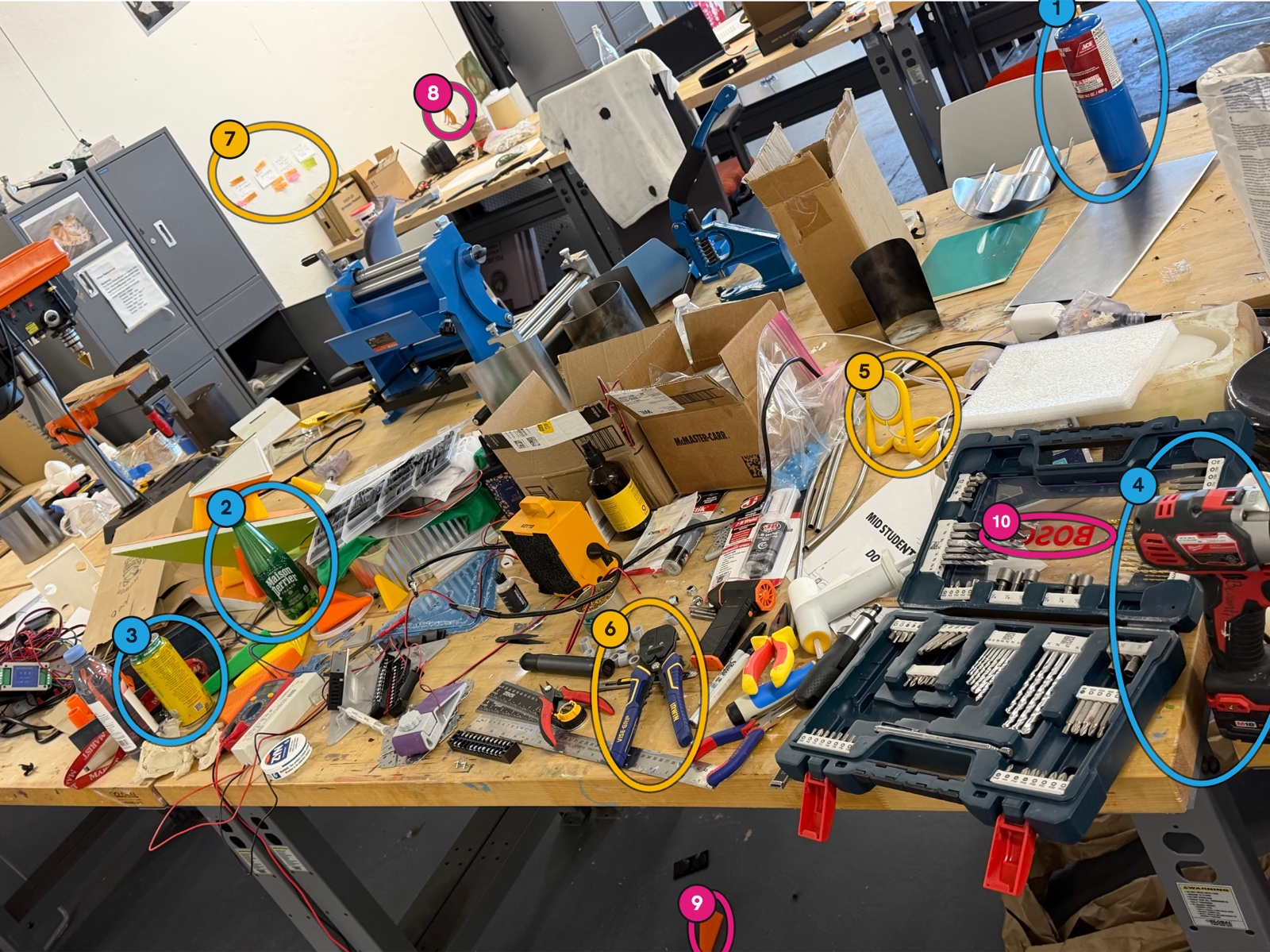 A photo of a busy studio workbench with ten finds circled and numbered: a blue gas tank, a green glass bottle, a yellow can, a red drill, a yellow phone stand, blue locking pliers, sticky notes, a tiny orange dog on the wall, an orange wedge on the floor and upside down lettering.